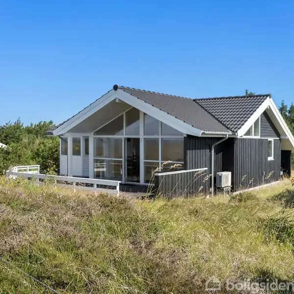 Et sort sommerhus med gråt tag står stille midt på en åben græsmark. I baggrunden ses kystlinjen og havet, omgivet af frodig vegetation og løvtræer.