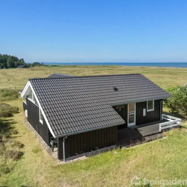 Et sommerhus med sort tag og hvide detaljer står omgivet af grønt græsland. I baggrunden ses en kystlinje og havet under en klar blå himmel.