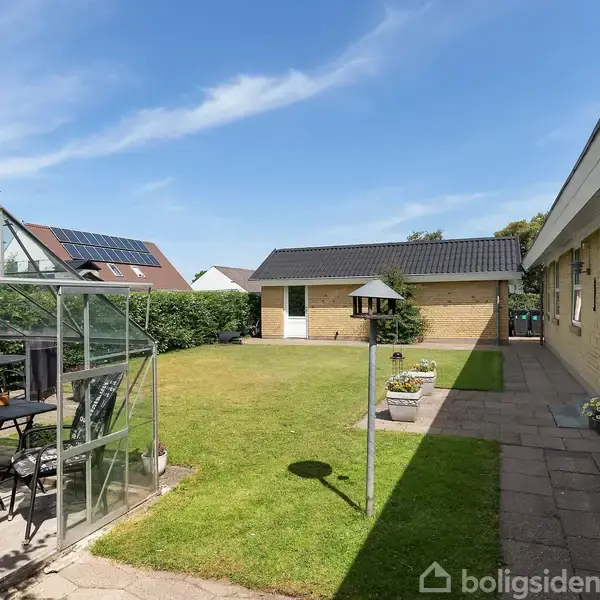 Et drivhus med sorte møbler står i en lys have ved siden af et gult murstenshus. Græsplænen er omkranset af hække, og der ses enkelte blomsterkrukker.