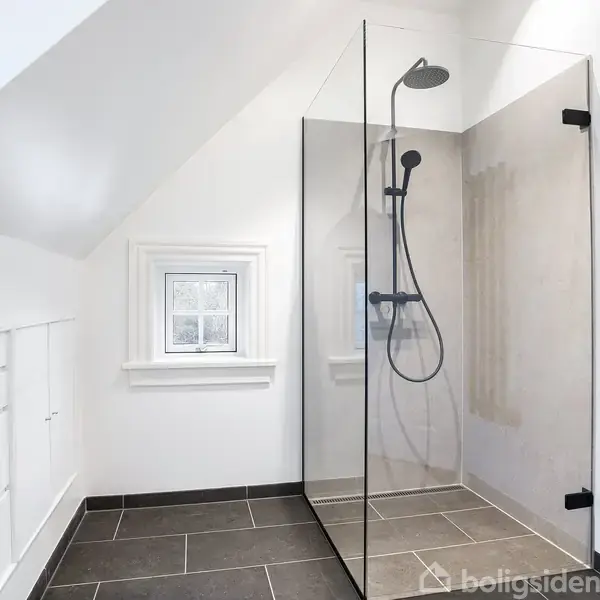 Et moderne bruseområde med glasvæg står i et badeværelse med skrå loft. Fliserne er mørke på gulvet, og væggene er hvide. Der er en lille vindueskarm på væggen.