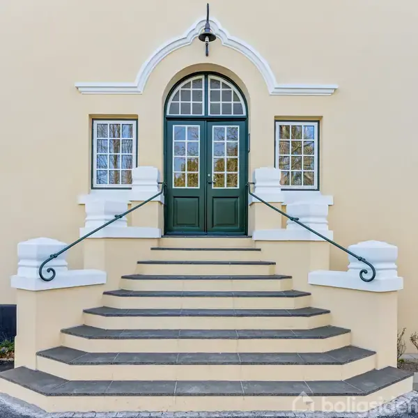 Trappe fører op til grøn dør med bueformet vindue, flankeret af symmetriske hvide vinduer. Beige bygning i baggrunden, med sorte tilføjelser og små planter ved siden af trappen.