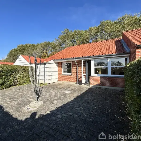 Rød murstenshus med gavl og rødt tegltag i solbeskinnet gårdhave omgivet af hæk. Foran huset står en enkelt, bladløs træstamme. Himlen er klar blå.