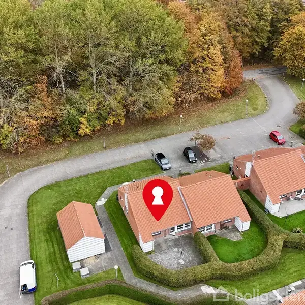 Hus markeret med rød nål ses fra oven, omgivet af frodige grønne haver, med en vej og parkerede biler til venstre. Baggrunden viser en tæt skov med efterårsfarver.