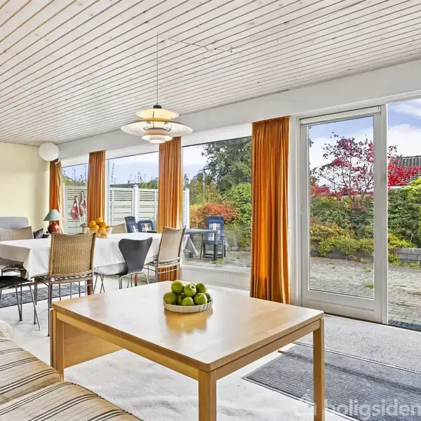 Stue med sofagruppe og spisebord ved siden af store vinduer. Franske døre åbner til en terrasse omgivet af have. Elegant loftslampe hænger over bordet.