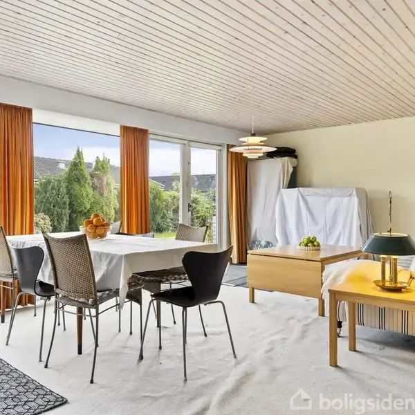 Spisebord med seks stole står i en stue med hvide vægge og loft. Store vinduer med orange gardiner viser en have med buske og træer udenfor.