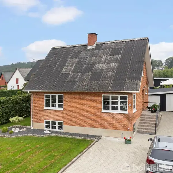 Et rødt murstenshus med sort tag står ved siden af en garage. Bilen parkeret i indkørslen. Grønne buske og træer omkranser området, mens en flagstang står i græsplænen.