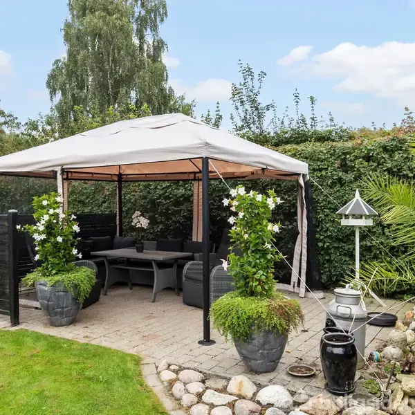 Pavillon står på flisebelagt område, omgivet af grønne planter og møbleret med bord og sofaer. Terrasse afgrænset af lav sort væg, placeret i grøn have.