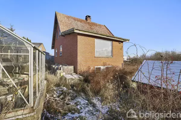 Murstenshus med skråt tag står i forsømt have med vildtvoksende planter og snepletter; til venstre ses et drivhus. Skyfri himmel i baggrunden giver stedet et øde udtryk.