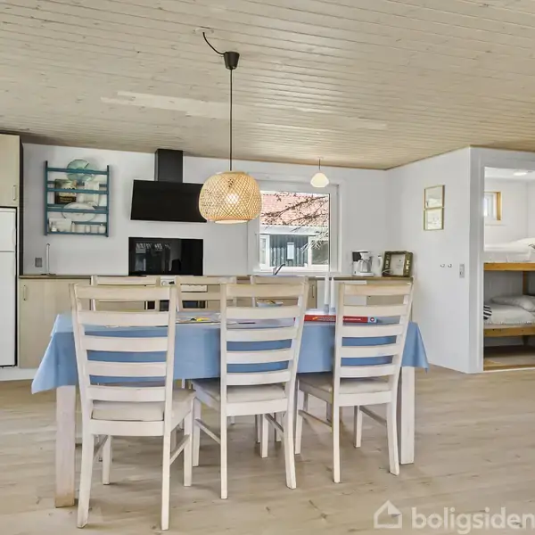 Spisebord med seks stole står i et lyst køkken-alrum. Bordet er dækket med en lys blå dug. Der er et ildsted til højre. En lampe hænger over bordet.