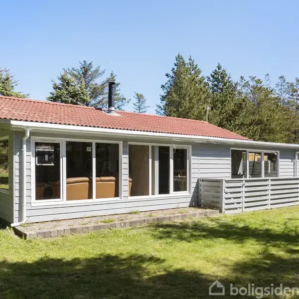 Et sommerhus med rødt tegltag og store vinduer står i en solrig have omgivet af træer. Græsplænen er nyklippet, og der er en terrasse foran huset.