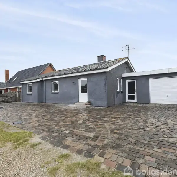 Grå bungalow med fladt tag, garagedør og flisebelagt forhave; placeret i et roligt villakvarter med lignende huse.