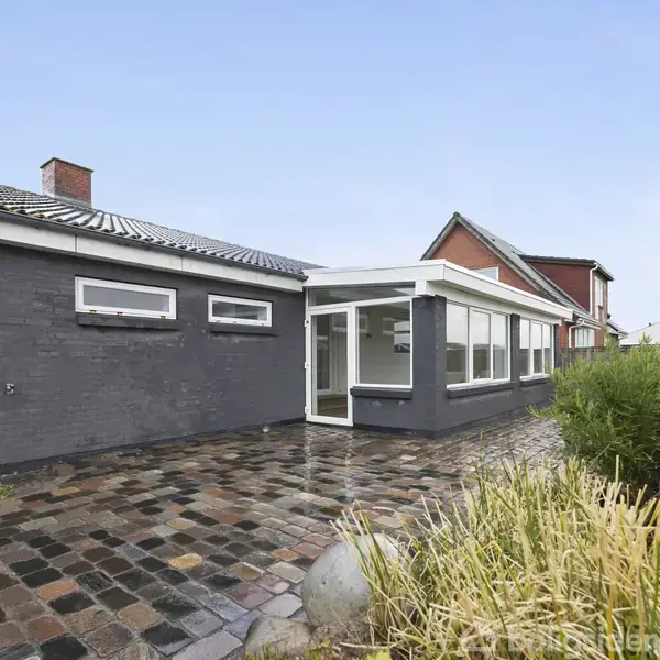 Et hus med grå murstensvægge og flere vinduer ligger ved en brostensbelagt gårdhave. Der er en tillukket veranda i hvid glasramme. Buske omgiver gårdhaven.
