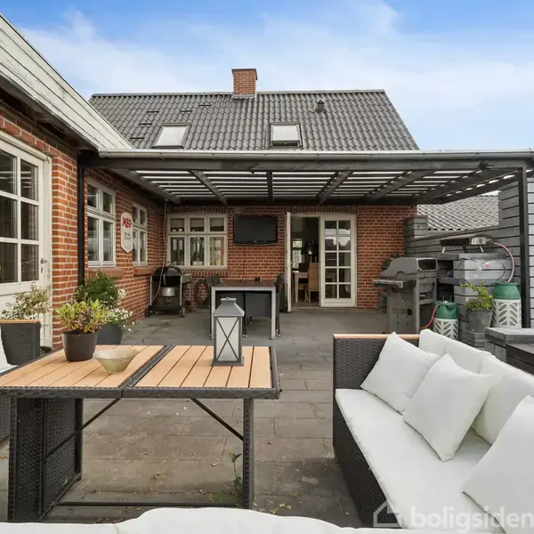 Terrassemøbler står på en stenterrasse foran en rød murstensbygning med vinduer og en glasdør. Et halvtag beskytter mod solen. Planter og grill ses i baggrunden.