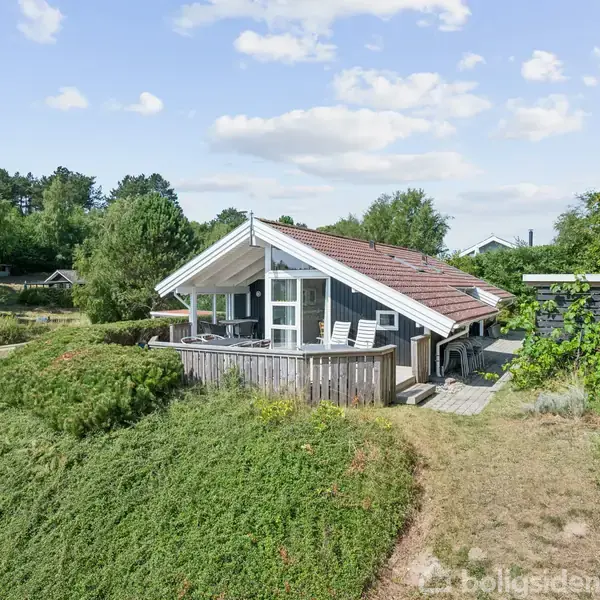 Et træhus med rødt tag står på en lille bakke. Foran huset er en terrasse med stole. Omgivet af grønt landskab og spredte træer under en blå himmel med skyer.