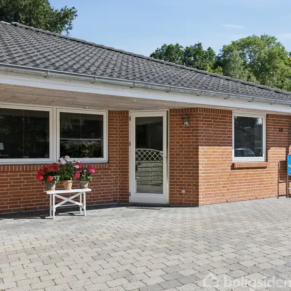 Rød murstensbungalow med gråt tegltag står på flisebelagt gårdsplads. Bord med blomster ved vinduet. Omkranset af grønne træer og hegn i baggrunden.