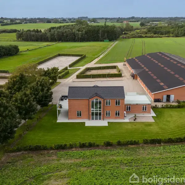 Murstenshus set forfra, omgivet af græsplæne og træer, ved siden af en stor lade. Baggrund præget af åbne marker og skovbryn under en klar himmel.