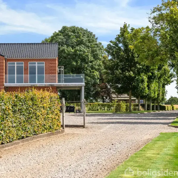 En rød murstensbygning med store vinduer står ved siden af en grusvej omgivet af grønne træer og hække, under en lys blå himmel med få skyer.