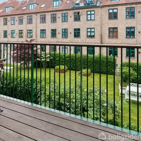 En gang med et trægulv, hvor flere jakker hænger på kroge. Et rødt spejl hænger på væggen. En åben dør fører til et køkken med ovn og emhætte.