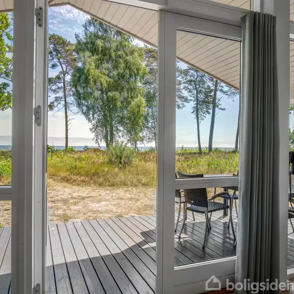Døre åbner ud til en terrasse med udsigt over en græsplæne og træer. Terrassebord og -stole står udenfor. Skoven og himlen ses i baggrunden.