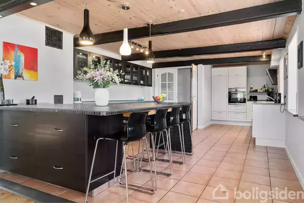 Et moderne køkken med en stor sort køkkenø og sorte barstole omkring. Over øen hænger lamper. Køkkenet har skabe, hvidevarer og en farverig vægdekoration.