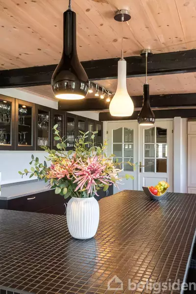 Vase med blomster står på sort flisebord under tre hængelamper i et køkken. Bagvaske hvide skabslåger og glaslåger med glas og porcelæn. Skål med frugt står ved siden af.