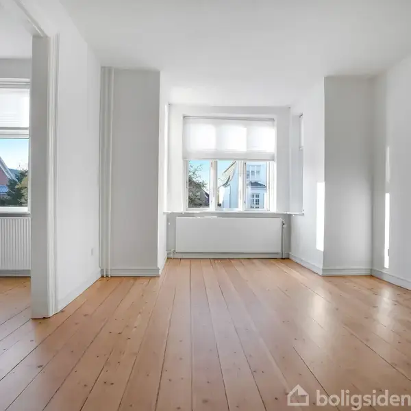 Et lyst, tomt værelse med trægulve og hvide vægge. Store vinduer slipper sollys ind, og en radiator er placeret under vindueskarmen. Vinduerne viser et udsyn til nabohusene.