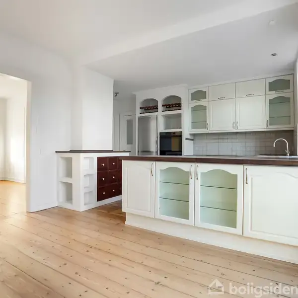 Køkken med hvide skabe og glaspaneler, træbordplade, og indbygget ovn. Åbent ind i stue med trægulv og stort vindue, hvilket giver et lyst og åbent rum.