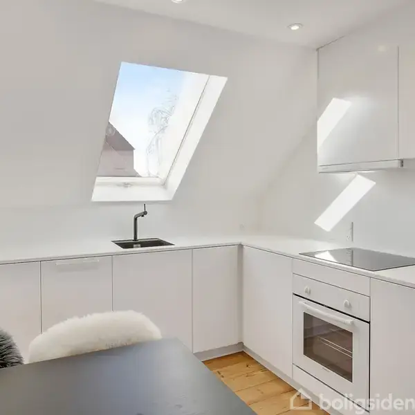 Et moderne køkken har hvide skabe og en indbygget ovn. Et stort, skråtstillet ovenlysvindue lyser rummet op. Et spisebord med pelstæppe ses i forgrunden.