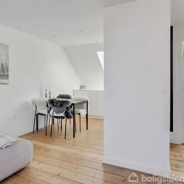 En stue med en grå sofa til venstre, et spisebord med fire stole i midten og et stort foto af en surfer på væggen. Lyse trægulve og hvidmalede vægge.