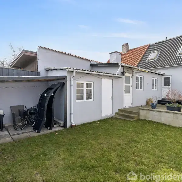 Et hvidt hus med flere tilbygninger, cykler under halvtag, og havemøbler på terrasse. Omkringliggende græsplæne med blomsterbede. Klar blå himmel i baggrunden. Ingen tekst i billedet.