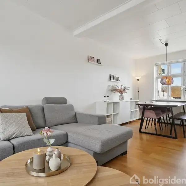 Grå sofa med puder står i en stue ved et rundt sofabord med lys og dekorationer. I baggrunden ses en spisestue med bord og stole, lyse vægge og trægulv.