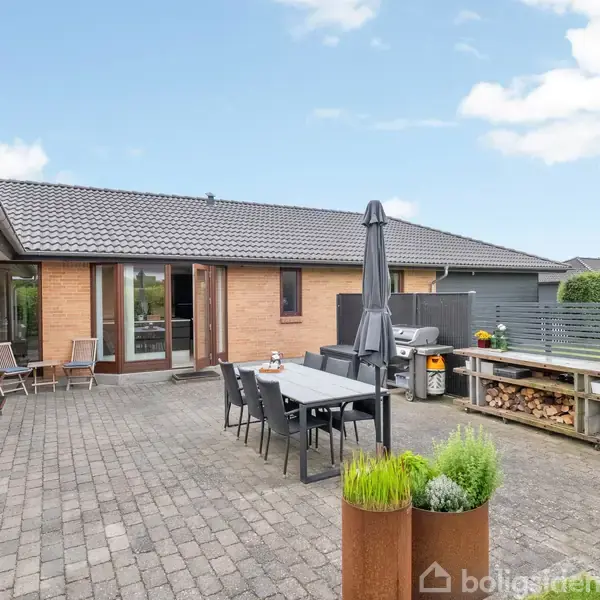 Et moderne køkken med en stor køkkenø, hvorpå der står en plante og en kurv med krydderier; åbne døre leder ud til en terrasse, og spisebordsområde ses i baggrunden.
