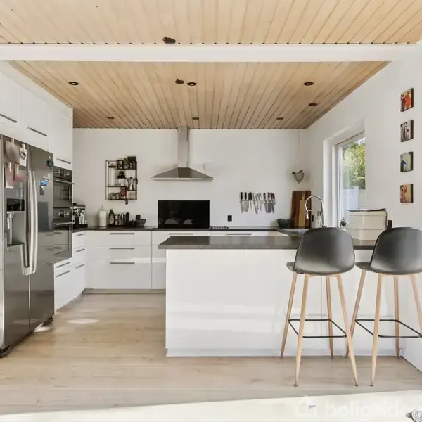 Køkken med hvidt køkkenskab, moderne køleskab og to høje stole langs en køkkenø. Træloft og store vinduer skaber en lys og åben atmosfære. Ingen tekst i billedet.