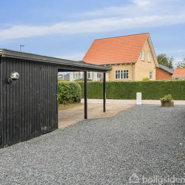 En sort garage står stille ved siden af et hus med rødt tag i grusbelagt indkørsel, omgivet af grønne buske og blå himmel.
