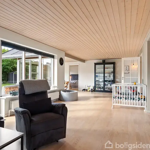 En stue har en lænestol, et sofabord, og et legeområde med et indhegnet legetøjsgitter. Store vinduer viser en terrasse og have udenfor, mens lyse trævægge og loft giver et åbent rum.