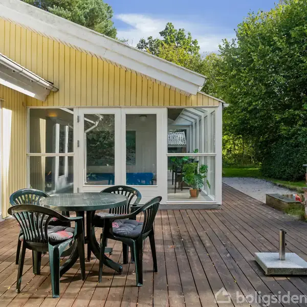 Rundt havebord med sorte stole står på en træterrasse. Omkring bordet er en gul husfacade med glasdøre og vinduer samt en frodig have i baggrunden.