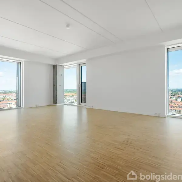 Et tomt rum med trægulv og hvide vægge, har to store vinduer med udsigt over byens skyline i dagslys. Ingen tekst er synlig i billedet bortset fra et logo.