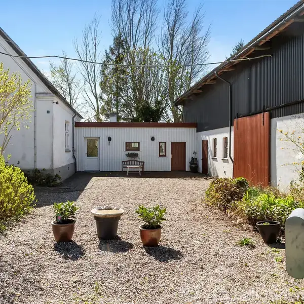 Gårdsplads omgivet af to bygninger, med grusbelægning og blomsterkrukker. En bænk står ved en væg i midten, omringet af grønne buske og en blå himmel.