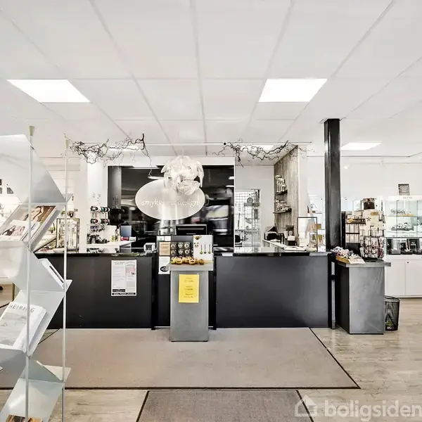 Butikinteriør med skranke i midten omgivet af hylder med varer. Diverse dekorationer hænger fra loftet. Der er en stor lampe over disken, og miljøet er lyst og rummeligt.