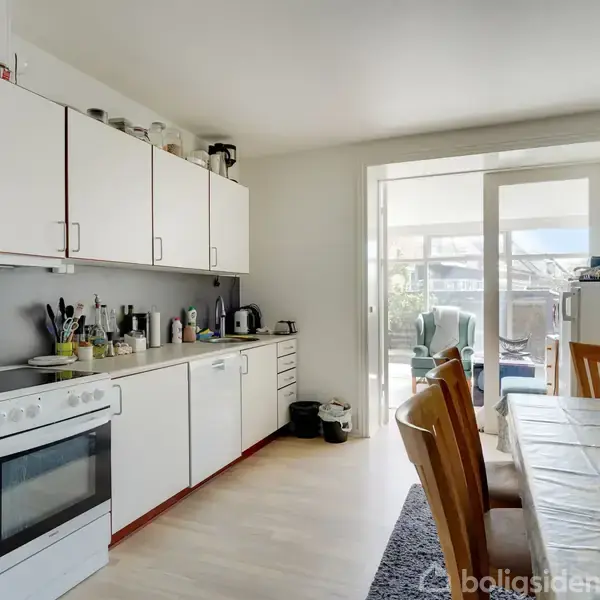 Et køkken med hvide skabe, en komfur og bordplade fyldt med køkkenredskaber. I forgrunden står et bord med træstole og en vase med pink tulipaner. I baggrunden ses en lys terrasse.