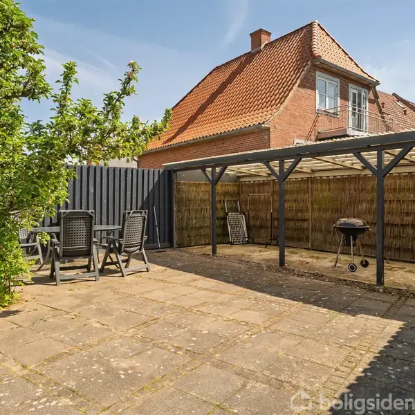 En flisebelagt gårdhave med havemøbler og en grill står under en pergola. Et træ er i nærheden, og i baggrunden ses et rødt murstenshus med tegltag.