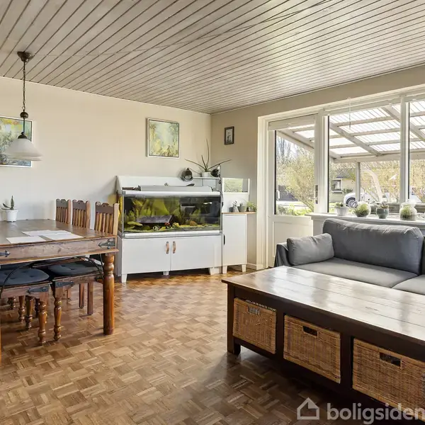 Spisestue med træbord og stole, et akvarium på et hvidt skab, grå sofagruppe og sofabord med kurve. Beliggende ved store vinduer mod overdækket terrasse og have.