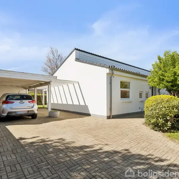 En hvid bil holder parkeret i en carport ved et hvidt hus. Foran huset er en flisebelagt indkørsel og en velplejet have med træer og buske under en klar blå himmel.