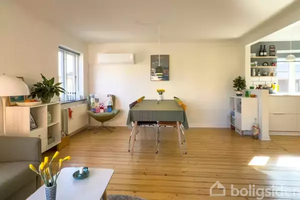 En spisestue med et bord dækket med en dug og fire stole. Rummet er lyst med træplankegulv, reoler ved væggen og dekorationer, inklusive planter og en lampe.