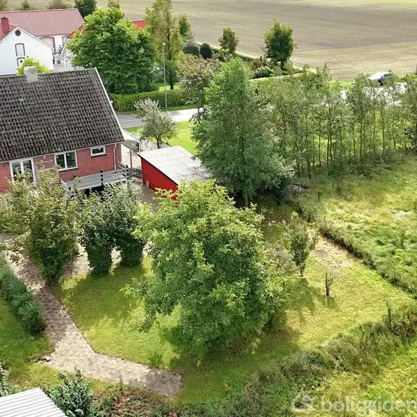Et rødt hus med skråt tag står i en grøn have med træer og buske. En sti fører fra huset gennem haven. Landskabet omkring er landligt med marker.
