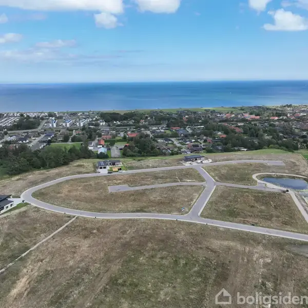 En nyudviklet byggeplads med vejnet strækker sig over en mark tæt på en kystby, omringet af træer og byggede huse. I baggrunden ses havet under en klar blå himmel.