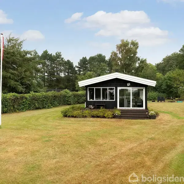 Et sort sommerhus står midt på en nyslået græsplæne. Ved siden af er der en flagstang. Baggrunden består af høje træer og tykkere buskads under en skyet himmel.