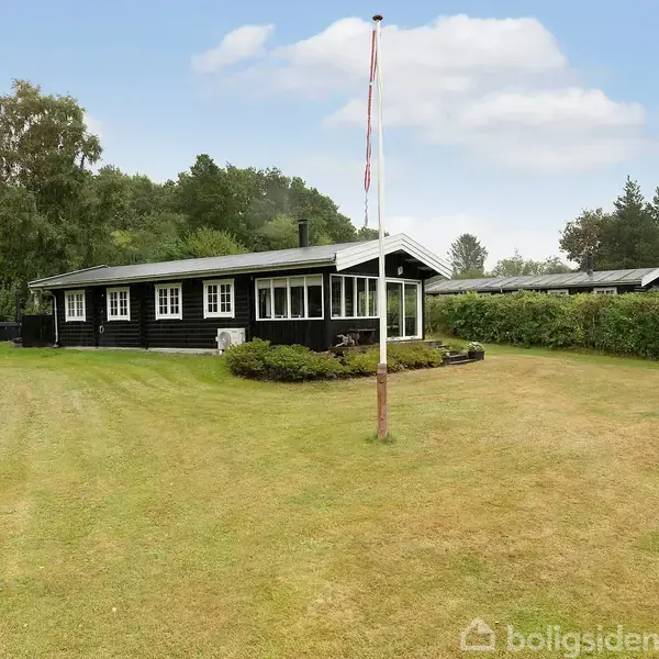 Et sort sommerhus med hvide vinduer står på en stor, velplejet græsplæne omgivet af buske og træer. Der er en flagstang foran huset under en skyfri himmel.