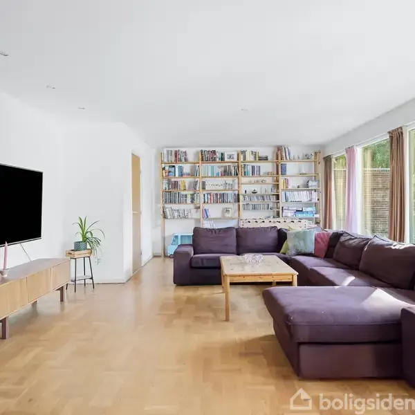 Stor, lilla hjørnesofa står i en stue med trægulv. Til venstre hænger et TV på væggen over et lavt møbel. Bagerst ses en fyldt bogreol. Store vinduer med gardiner.