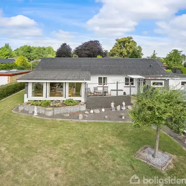 Huset har en bred terrasse med stenstatuer, omkranset af en velplejet græsplæne og træer. Beliggende i et boligområde med naboer og blå himmel i baggrunden.